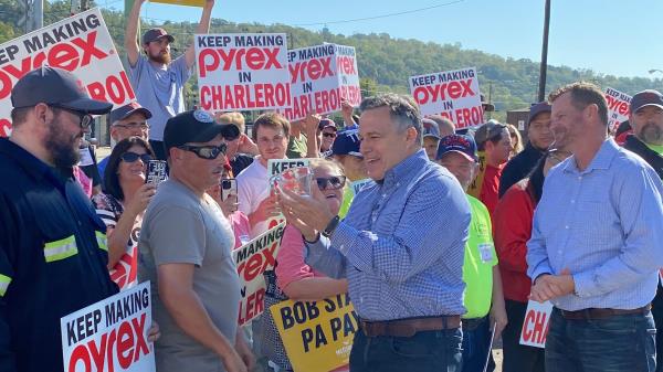 Dave McCormick holds up a Pyrex measuring cup, given to him by a worker. Signs reading "KEEP MAKING PYREX IN CHARLEROI" surround.