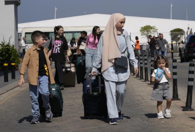 A woman with a religihous headcovering walks with luggage, with a boy and a girl on either side of her. In the background are more people wheeling luggage.