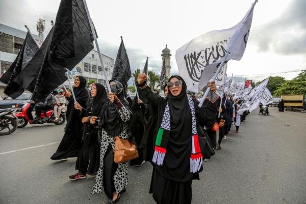 A group of Muslim women wearing head-to-toe black garb wave black and white flags as they march in formation down a street.