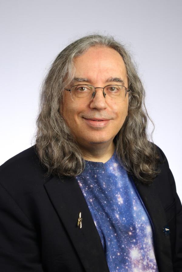 Live Space Race: Man with long gray hair smiles in a portrait. He is wearing a star-studded shirt and a dark blazer.